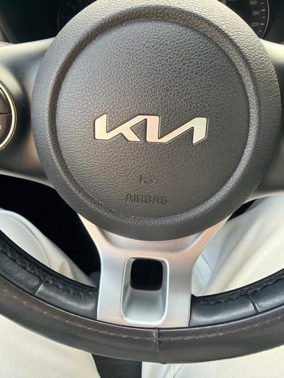 The image shows the steering wheel of a Kia car. The steering wheel has a black leather rim and a center hub with the Kia logo. The logo is silver and stylized. The word "AIRBAG" is printed on the hub next to the logo. A silver-colored spoke extends from the hub to the rim. The image does not provide any information to identify a location in Murmansk, Russia, or any other specific details about people, actions, or the environment.