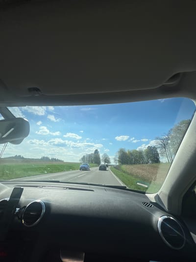 The image, taken from inside a vehicle in Muttensweiler, Germany, captures a view through the windshield on a bright, sunny day. The perspective is looking forward from the passenger or driver's seat.

Inside the car, a grey sun visor is visible on the left, with the A-pillar framing the view. The dark, textured dashboard occupies the lower right and central foreground. Visible dashboard elements include circular air vents with metallic trim on the right side, rectangular vents closer to the center, and a speaker grille near the right A-pillar. A partial view of the rear-view mirror is present in the lower center.

Outside the vehicle, a road stretches into the distance, with at least two cars visible ahead. The sky is a clear, vibrant blue with scattered white cumulus clouds. To the left and right of the road, the landscape consists of green fields and scattered trees, some with bare branches and others showing green foliage, suggesting an early spring or late autumn setting. Strong sunlight causes reflections on the windshield and dashboard.

A green, circular sticker is affixed to the lower left portion of the windshield, but any text or symbols on it are not clearly legible due to distance and resolution. The overall scene depicts a calm drive through a scenic, rural or semi-rural area under fair weather conditions.
