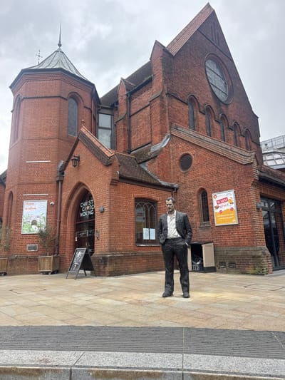 This image captures the exterior of Christ Church Woking, a red-brick ecclesiastical building located in Woking, United Kingdom, under an overcast sky. The prominent structure features a large main section with a high-pitched, tiled roof and a significant circular stained-glass window at its apex. To the left, a multi-sided tower with arched windows culminates in a conical metallic spire topped with a finial and a weather vane incorporating a cross.
A dark-colored, life-sized statue of a man stands on the light-colored paved forecourt directly in front of the church. The man is depicted wearing a suit jacket, an open-collared shirt, and trousers, with his hands placed in his pockets. The statue has realistic features and is positioned facing slightly right from the viewer's perspective.
The church's entrance area, beneath an arched red-brick canopy, features glass doors displaying "CHRIST CHURCH WOKING." To the left of the entrance, a poster titled "Hope is alive" is mounted on the wall. To the right of the entrance, another poster advertises a "WARM SPACE" offering "Refreshments and a warm welcome! free," attributed to Christ Church. An A-frame chalkboard sign, partially visible near the entrance, advertises a "Coffee Shop."
The immediate ground consists of uniformly laid square paving slabs in a light tan hue. A dark grey, horizontally ridged tactile paving strip is visible in the lower foreground, bordering a darker grey curb. Wooden planters containing small, bare-branched trees are situated along the church wall to the left of the entrance. No human subjects or active interactions are visible in the scene.