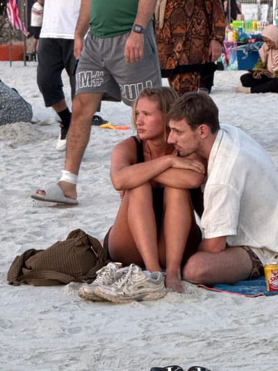 The image depicts a crowded beach scene at Cenang Beach, Malaysia. In the foreground, a blonde-haired female is seated directly on the sand, wearing a dark top and shorts, with her knees bent and arms crossed over her chest. Her gaze is directed towards the left and slightly upward. To her right, a brown-haired male is seated on a blue towel, wearing a light-colored, short-sleeved shirt and patterned swim shorts. His gaze also points towards the left. A pair of white athletic shoes rest on the sand in front of the female, next to a brown woven bag. A red and yellow beverage can is visible next to the male's right leg on the towel.

In the midground, a male stands to the left of the seated pair. He wears a green t-shirt, grey athletic shorts displaying "M#" and "RUN," and a black digital watch on his left wrist. His right foot wears a white slide sandal, with a white wrap around his right ankle. His left foot, partially visible, has a black sock and a white sneaker. Behind him, another male's legs are visible in dark shorts and black sneakers. Further back, a person wearing a brown and orange patterned garment stands. On the far right, a female in a light-colored hijab is seated on the sand, looking down at a small object. The background contains additional indistinct figures and colorful items, possibly vendor stalls. A section of an American flag is visible on the far left. The ground consists of light-colored sand, illuminated by soft, ambient light.
