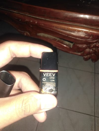 A human hand, with visible skin texture, holds a black, rectangular VEEV ONE vaping pod in a vertical orientation. The pod displays "VEEV one" and "3.5%" nicotine strength. Partially legible text below indicates "Philip Morris Products S.A., Quai Jeanrenaud 3, 2000 Neuchâtel, Switzerland." The lower portion of the pod shows a significant accumulation of dark, viscous residue. To the immediate left of the hand, a dark, elongated, hollow object, consistent with a component of a VEEV ONE vaping device, is partially visible. The background features a dark brown, carved wooden surface, likely furniture, positioned diagonally across the upper frame. Below the hand, a light gray tiled floor with distinct grout lines is visible. The scene is dimly lit, suggesting an indoor environment. The specific location within Uccle - Ukkel, Belgium, cannot be visually confirmed from the image but is presumed to be the setting as per the inquiry.