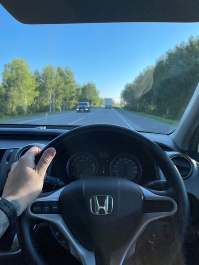 The image shows a first-person perspective from inside a car, driving on a paved road lined with trees. The driver's left hand, which has visible tattoos, is on the steering wheel. The dashboard displays a speedometer reading approximately 60 km/h, and various buttons for the audio system are visible on the steering wheel. The sky is clear and blue. In the distance, another vehicle, a truck, and a dark car are visible on the road. The scene appears to be a rural or suburban area, possibly during daylight hours with good visibility. There is no indication of the city of Balta, Russia.
