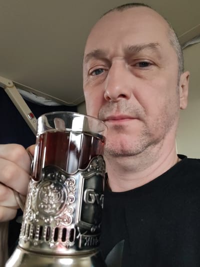 A middle-aged man with short, dark, receding hair, blue eyes, and light stubble on his face is depicted in a close-up selfie. He wears a plain black crew-neck shirt and has a neutral or slightly serious expression.
He is holding a clear glass filled with a dark liquid, likely tea, encased in an ornate silver-colored *podstakannik* (metal glass holder). The *podstakannik* features intricate embossed designs, including a prominent double-headed eagle emblem, commonly associated with Russia. Faint Cyrillic text is visible on its side, reading "СГУП" and "РЖД", with "РЖД" being the acronym for Russian Railways (Российские железные дороги).
The setting appears to be the interior of a train carriage or an airplane cabin, suggested by the light-colored overhead panel and a folded, beige-colored tray table arm with a small red button and an icon. The man is holding the glass up towards the camera, seemingly taking a picture while seated. The location context is Бордуки, Russia, which aligns with the Russian Railways branding on the *podstakannik*.