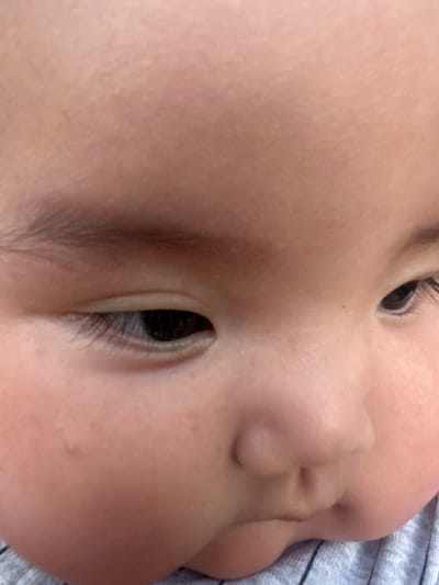 The image is a close-up, angled shot of a baby's face. The baby's left eye is partially visible, with long eyelashes and a dark iris. The baby's cheek is plump and soft, with a few scattered freckles. The skin tone is fair and appears healthy. The baby is wearing a grey, striped garment, likely a shirt. The background is blurred and out of focus, suggesting a shallow depth of field. The lighting is soft and natural, casting a gentle glow on the baby's skin. The image conveys a sense of intimacy and tenderness, capturing a fleeting moment of childhood. The provided metadata about Manas, Kyrgyzstan is irrelevant to the visual content of the image.