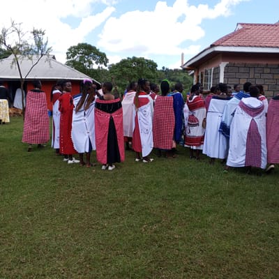 A group of people, primarily women, are gathered on a grassy lawn near two buildings. The buildings appear to be single-story structures. The women are wearing traditional clothing, including red and white patterned wraps. The scene is lit by daylight, and there are trees and a slightly cloudy sky in the background. The location appears to be Keyian ward, Kenya.