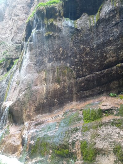 The image shows a natural rock face with a waterfall, covered in moss and vegetation. The rock is layered and appears to be a cliff face. Water is flowing down the rock in several places, creating small cascades and streaks. The ground at the base of the cliff is wet and appears to be part of a river or stream. The overall scene is one of natural beauty and is likely a scenic overlook or a natural park. The location is identified as Khushto-Syrt, Russia. There are no people or animals visible in the image.