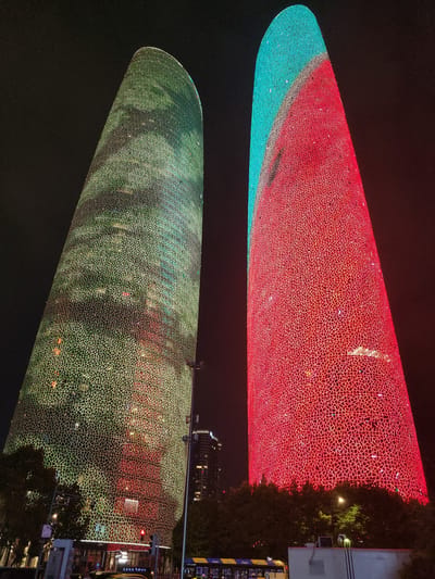 Two illuminated skyscrapers dominate the night sky in Wuhou District, China. The skyscraper on the left is covered in a green, illuminated geometric pattern, while the skyscraper on the right displays a red and turquoise pattern. The buildings are tall and curved, tapering towards the top. The base of the buildings is surrounded by trees, and a street with traffic lights and vehicles is visible at the bottom of the frame. In the background, other buildings are also lit up. The overall scene depicts a vibrant urban night landscape.
