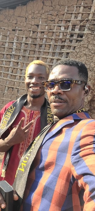 An outdoor portrait depicts two adult males positioned in front of a wattle and daub wall, characteristic of rural architecture, located in Alayi, Nigeria. The subject on the left, a male with blond hair, smiles broadly, displaying teeth, and makes a 'V' gesture with his left hand. He wears a maroon long-sleeved shirt with an embroidered neckline and a dark sash with reversed yellow text diagonally across his chest. The subject on the right, an older male, wears dark, square-framed sunglasses, a blue and orange striped jacket or shirt, and a dark sash with reversed yellow text across his chest. He has a mustache, beard, and a black earbud in his right ear. He holds a dark, rectangular object in his left hand. Both individuals are looking towards the camera.