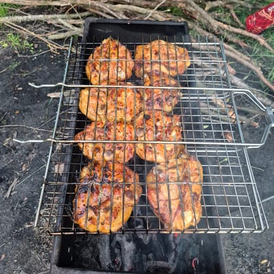 The image shows a close-up view of chicken pieces grilling on a metal rack over hot coals. The chicken is golden brown with char marks, indicating it is cooked. The grilling rack is placed on top of a dark metal barbecue. The background consists of dry branches and some green grass, suggesting an outdoor setting. The ground appears to be dirt and ash. There is a red object with white lettering visible in the upper right corner of the image, but its identity is unclear. The scene is consistent with a barbecue event in Chmyrivka, Ukraine, given the rustic, natural surroundings.