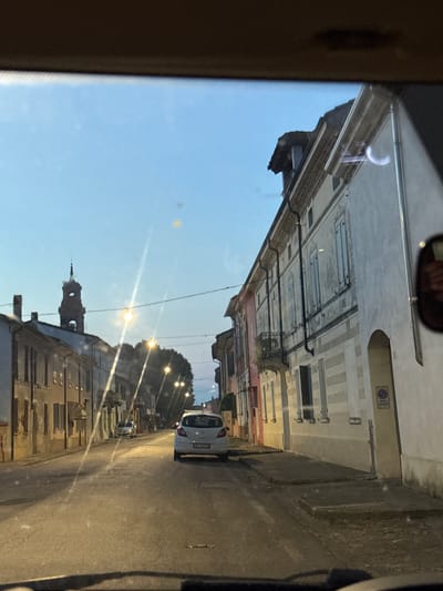 The image depicts a street scene in Villa Pasquali, Italy, captured from the perspective of someone inside a vehicle. The time appears to be dusk or early evening, as the sky is a pale blue with a gradient of lighter hues near the horizon. Streetlights are illuminated, casting long shadows on the asphalt road.

The street is lined with buildings on both sides. On the right, a row of multi-story buildings exhibits a slightly aged facade with shutters on the windows. One building features a prominent archway leading to an unseen area, and a sign with Italian text is visible near it. The building on the far right shows a rounded reflection on its surface. On the left, buildings are also visible, appearing to be residential or commercial. In the distance, a clock tower with an ornate steeple rises above the buildings, its silhouette sharp against the sky.

A white car is stationary in the middle of the road, facing away from the viewer. Another silver car is parked further down the street on the left. The road surface is asphalt with visible lane markings. Power lines and streetlights are strung across the street. The overall impression is of a quiet residential or town center street during the evening transition.