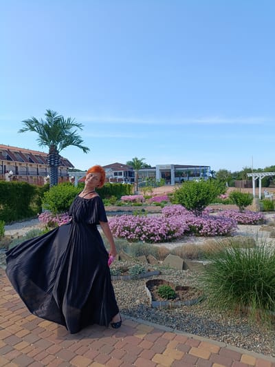 A woman with bright orange hair and a black, flowing dress stands in a garden under a clear blue sky. She is posing with her right arm extended upwards and her body tilted to the left, her dress billowing out around her. The garden features a variety of plants, including a palm tree, bushes, and beds of small pink flowers. In the background, there are buildings with terracotta roofs and a white structure with a canopy. The ground in the foreground is paved with reddish and yellow bricks. The scene appears to be set during the daytime with sunny weather. The woman's expression is cheerful. The text "Kris Hall" is visible on a building in the background. The location is Za Rodinu, Russia.