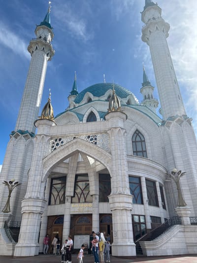The Kul Sharif Mosque in Russia is depicted under a bright, clear blue sky with sparse white clouds. The grand mosque features an ornate white stone facade with intricate carvings, geometric patterns, and numerous arches. Two prominent, towering minarets with decorative balconies and turquoise-tipped spires flank the central structure. A large, ribbed turquoise-blue main dome and several smaller matching domes adorn the roof, each topped with a golden finial.
The building exhibits various window designs, some with dark frames and intricate internal patterns. Ornate, branching light fixtures with spherical lamps are visible on the exterior walls. Near the mosque's entrance, several people, including adults and children, are gathered, indicating a public visitation or tourist activity. Above an entrance, Cyrillic text clearly reads "Кол Шәриф" (Kul Sharif) and "Мәчете" (Mosque), identifying the building. The scene captures the mosque's magnificent architecture on a sunny day in Russia.