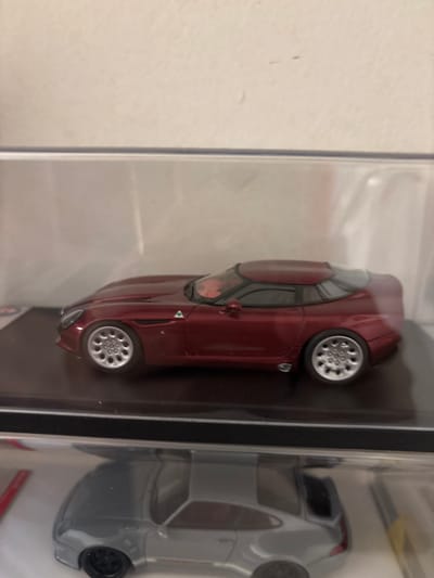 The image displays two clear plastic display cases, stacked vertically, against a plain light-colored wall. The upper display case contains a dark metallic red/maroon die-cast model car, presented in a side profile facing left. This model features silver multi-spoke wheels, a coupe body with a distinct roofline, and a visible Alfa Romeo emblem on its side panel behind the front wheel. Faint red interior details are visible through the windows. The lower display case, partially visible, contains a silver/light grey die-cast model car, also in side profile, featuring a prominent rear spoiler and black wheels. No people or dynamic actions are depicted. The environment is an indoor setting. The specific geographical location within the city of Kajang Municipal Council, Malaysia, cannot be determined from the visual information provided in the image.
