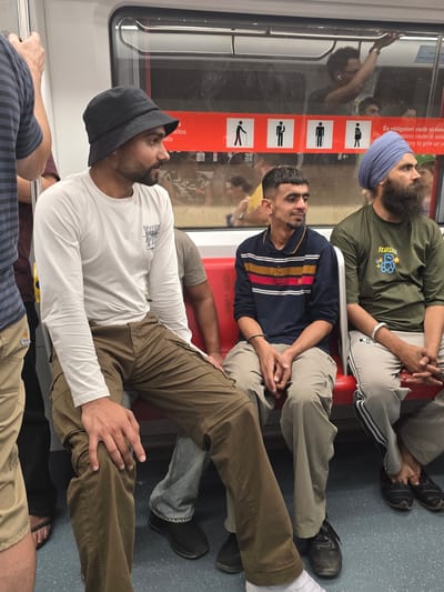 The image shows the interior of a public transport vehicle, possibly a subway or train, with several individuals seated. The setting appears to be in Cornellà De Llobregat, Spain, as indicated by signage in Spanish visible through the window.
In the foreground, three men are prominently featured. The man on the left is wearing a white long-sleeved shirt and brown cargo pants, with a black bucket hat. He is looking to his right. To his right, a man in a dark blue polo shirt with horizontal stripes of red, yellow, and white is seated, looking to his left. He has short, dark hair and is wearing light brown pants. On the far right, a man wearing a green t-shirt and a blue turban is seated, looking towards the right. His t-shirt has a graphic of a teddy bear and the word "FEARLESS" on it. He has a long, dark beard.
The seating appears to be red upholstered benches. Behind the seated individuals, there is a window revealing the outside, or possibly another part of the vehicle, where other passengers are visible. A red band with pictograms of people, including one with a cane and a pregnant woman, runs horizontally across the interior of the vehicle. This band is likely indicating priority seating or rules for passengers. The floor is a speckled gray material. The overall environment suggests a functional, urban public transportation setting.