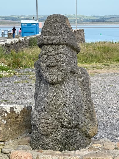 The image depicts a stone sculpture of a figure wearing a hat, situated outdoors in a coastal environment. The sculpture is made of a dark, porous rock and has a roughly carved, anthropomorphic appearance with discernible facial features and a rounded body. In the background, there are two people walking on a paved path next to a bright blue portable toilet. Beyond the path, a body of water is visible, with rolling green hills and a clear sky in the distance. A green buoy floats in the water. The ground in the foreground and around the sculpture is covered in gravel and some larger, rough stones, suggesting an unpaved or partially developed area. The overall scene appears to be in a public outdoor space, possibly a park or a scenic viewpoint. The city of Terhillion, Ireland is not discernible from the image itself, and no specific landmarks are present to confirm this location.