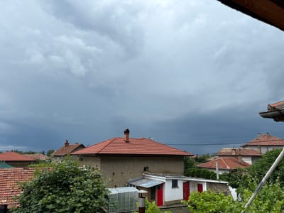 The image presents an overcast daytime view of a village, identified as Hadzhidimitrovo, Bulgaria, seen from an elevated perspective. The dominant feature is a vast, heavy, dark grey sky filled with dense, low-hanging clouds, strongly suggesting an impending storm or recent adverse weather.
Below the dramatic sky, a cluster of houses with characteristic terracotta red tile roofs is visible, interspersed with abundant green trees and bushes. The architecture includes buildings with stuccoed walls, some appearing in neutral tones like beige or grey. Several chimneys protrude from the rooftops, particularly on a prominent two-story house in the midground. This house features a large, simple facade and a well-maintained red tile roof. Adjacent to it, a smaller, white auxiliary building with two distinct red doors and a corrugated metal roof stands out. A greenhouse is also partially visible near the lower-left corner.
Lush green foliage, including what appears to be grapevines, fills the lower and mid-sections of the frame, obscuring some of the structures. Power lines stretch horizontally across the midground, connecting unseen poles. The scene is static, with no human activity or visible text present. The overall mood conveyed by the dark sky is one of quiet anticipation or a somber atmosphere.