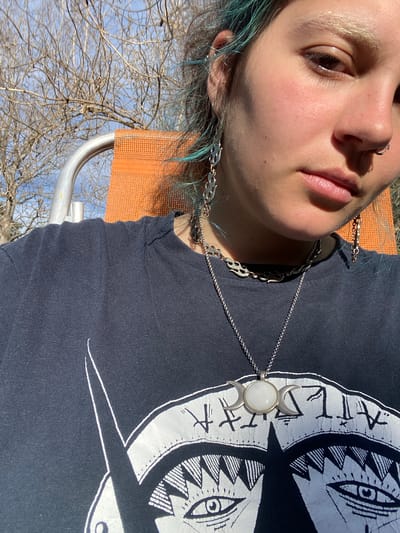 A close-up, chest-up, medium-angle shot shows a person with light skin and blue-tinted hair, facing forward and slightly to the right. They are wearing a dark t-shirt with a white graphic design featuring stylized eyes and symbols, and a silver necklace with a crescent moon pendant. The person also wears multiple silver earrings and a nose ring. The background consists of bare tree branches against a bright blue sky, and a sliver of an orange chair is visible on the left side of the frame. The lighting suggests it is daytime and sunny. The image does not provide information regarding the city of Durazno, Argentina.