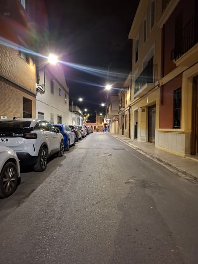 The image shows a narrow street in Xirivella, Spain, at night. The street is lined with parked cars on one side and buildings on both sides. Streetlights illuminate the scene, casting long shadows. The buildings appear to be residential, with multiple stories and a mix of architectural styles. The street surface is asphalt, showing signs of wear and tear. The overall impression is that of a quiet, residential neighborhood at night.