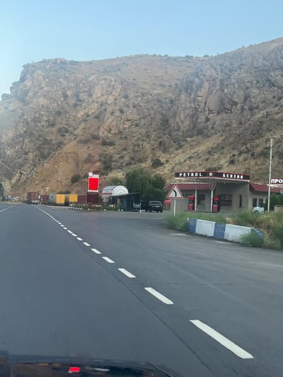 The image captures a roadside scene in the vicinity of Arpi, Armenia, characterized by a rocky mountain slope to the left and a gas station on the right. A paved road with white dashed lines dominates the foreground, suggesting a highway or major thoroughfare.
On the right side of the road, a PETROL station is visible, identified by a sign with "PETROL" in English and "БЕНЗИН" in Cyrillic script. The gas station has a covered canopy with lighting, and two fuel pumps are positioned beneath it. A white vehicle is parked to the right of the gas station. To the left of the gas station, a large digital display board shows a price of "210" in red illuminated digits. Adjacent to the digital display, several freight trucks are parked, their cargo containers varying in color, including red and yellow. Further back, a building with a curved roof is situated behind the gas station.
The environment is natural and somewhat arid, with sparse vegetation on the mountain slopes. The sky above is clear and a pale blue. The lighting suggests it is either early morning or late afternoon. There are no visible people in the frame. The perspective of the image indicates it was taken from a moving vehicle.