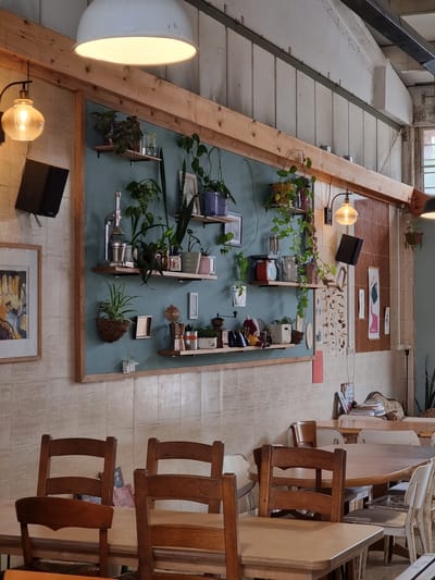 The image shows an interior space in Mitzpe Ramon, Israel. The viewpoint is directed upwards, emphasizing the vertical layout of the scene. A large wall-mounted display dominates the left side of the frame, showcasing an assortment of potted plants, small objects, and framed pictures arranged on a light blue surface with a wooden frame. The display appears to be an artistic installation or a decorative arrangement. Two lamps, one a large dome-shaped light and the other a smaller bulb lamp, illuminate the display. To the right, a series of wooden chairs and tables are stacked and arranged, suggesting a storage area or a workspace. The floor is tiled, and glimpses of an exterior window and architectural details are visible at the top of the frame. The overall atmosphere is that of a studio, workshop, or gallery setting.