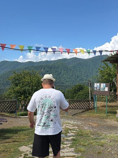 In Izmaylovka, Russia, a man wearing a white t-shirt with a dragon graphic and black shorts stands facing away from the camera. He is also wearing a light-colored fedora hat. The background shows a lush, green mountain range under a bright blue sky with scattered white clouds. Colorful triangular flags are strung across the top of the frame, and a wooden fence is visible in the middle ground. A wooden structure and a bulletin board are to the right of the man. The ground is a mix of gravel and stone pathways, with patches of grass.