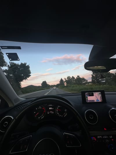 A first-person perspective inside a car shows the dashboard, steering wheel, and the view through the windshield. The car is driving on a road with trees and hills on either side. The sky is a mixture of blue and pink, indicating dusk or dawn. A navigation screen on the dashboard displays a map and route information. The car's speedometer shows a speed of 60 km/h. A temperature gauge reads 21.5 degrees Celsius. The location is the city of Vasilkovska mahala, Bulgaria. There are no people visible in the image.