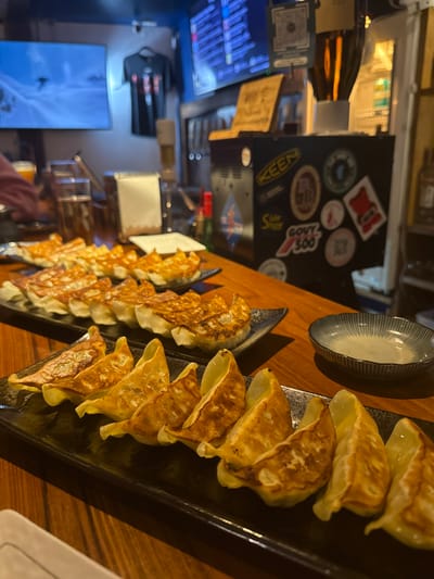 This image captures an interior view of Craftroom, a bar/restaurant located in Nozawaonsen, Japan, focusing on a wooden counter laden with food and drinks.
In the foreground, three black rectangular plates are arranged on the polished wooden counter, each presenting approximately ten pan-fried gyoza. The gyoza exhibit a golden-brown, crispy bottom surface. To the right of the foremost plate, a small, fluted ceramic bowl contains a light-colored dipping sauce.
In the midground, a person's left hand, partially visible on the far left, reaches towards a glass containing an amber beverage. Several other glasses, some holding similar amber liquid, are scattered along the counter. A white folded napkin or menu stands upright further back on the counter.
The background reveals a bar area with a predominantly blue wall. Two flat-screen televisions are mounted on the wall; the left screen displays a snowy mountain landscape with a skier, while the right screen shows what appears to be a digital menu or information board with Japanese text. A black t-shirt with a red design hangs on the wall between the two screens. Below the televisions, a black utility unit or cooler is heavily adorned with various stickers, including prominent "KEEN," "GOVY 500," and "Slide Show" decals. Multiple beer taps and various bottles are visible behind the counter. The overall lighting is dim, suggesting an evening setting, with localized illumination on the counter and bar.