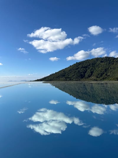 An abstract symmetrical view features a verdant hill juxtaposed with a clear blue sky adorned with scattered white clouds. The hill and sky appear mirrored along a central vertical axis. The clouds, resembling fluffy shapes, add texture to the sky. The hill is covered in dense green foliage, creating a sharp contrast with the blue expanse. A thin, light-colored strip runs along the base of the hill and the horizon, possibly a road.