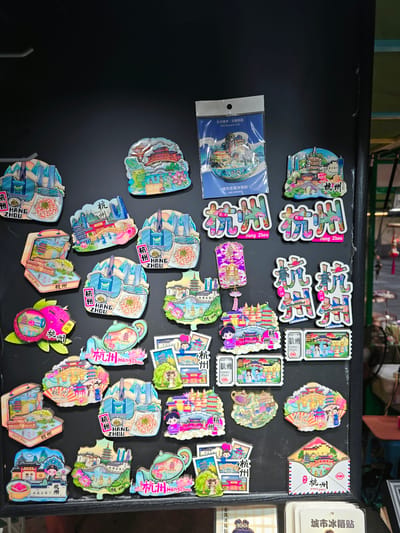 The image displays a collection of magnets, likely souvenirs, arranged on a dark, flat surface, possibly a wall or display board. The magnets feature colorful illustrations of scenic views and landmarks, predominantly related to Hangzhou, China. Many magnets are shaped like traditional Chinese buildings, pagodas, or fans, and some have the text "Hangzhou" written on them. There are also magnets with cartoon figures and traditional motifs like lotuses and teapots. The overall impression is of a retail display for tourist memorabilia. The scene appears to be indoors, with the dark surface suggesting a blackboard or a similar dark-colored material used for display. There are no people clearly visible in the image. While the specific location within Gongshu District is not explicitly stated, the items suggest a souvenir shop or market stall catering to tourists in that area of Hangzhou, China.