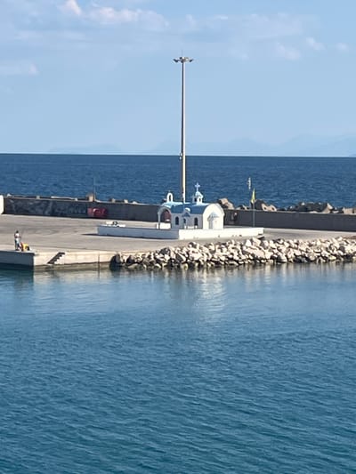 The image depicts a small, white chapel with a blue roof and a cross atop it, situated on a stone embankment by the sea. The chapel appears to be a landmark at a harbor or coastal area. In the background, a concrete wall lines the water's edge, and beyond that, a large, tall pole with lights stands prominently. The sea is a deep blue, with subtle waves, and a hazy line of distant mountains is visible on the horizon under a clear sky with some scattered clouds. In the foreground, on a concrete pier, a person is seen fishing, with a yellow object near them. The overall scene suggests a peaceful coastal location within the Municipal Unit of Kastro - Kyllini, Greece.