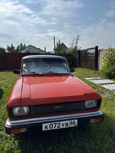 The image shows a red AZLH car parked on a grassy area in the city of Bogdanovich, Russia. The car is viewed from the front, with its hood and grille prominently displayed. The license plate reads "K072YB 66 RUS" with a Russian flag emblem. In the background, there are fences, trees, and houses, suggesting a residential or semi-rural setting. The sky is partly cloudy.