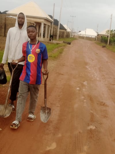 Two young males are standing on a dirt road in Russo, Nigeria. The male in the foreground is wearing a blue and red striped soccer jersey, grey pants, and white and black slide sandals. He is holding a shovel with a wooden handle and a metal blade. Around his neck, he wears a necklace with a medal featuring a yellow circular emblem with three yellow curved lines. The male in the background is wearing a grey hooded sweatshirt and dark pants. He is carrying a black plastic bag in his left hand and holding a shovel with a metal handle and blade in his right.

The setting appears to be a developing residential area with newly constructed houses lining the road. The houses have light-colored roofs and some have pillars at their entrances. The road is made of reddish-brown dirt and shows signs of recent rain with some muddy patches. Utility poles with wires are visible along the road. The sky is overcast, suggesting a cloudy or rainy day. The overall scene depicts two individuals possibly engaged in some form of construction or labor work, given the presence of shovels and the unfinished nature of the surroundings. The text "adidas" is visible on the slide sandals of the male in the foreground.