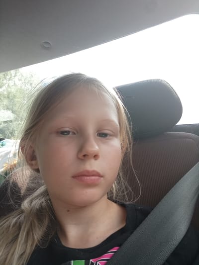 A young girl with long, blonde hair is shown in a selfie taken from inside a car. She is seated in the back, with a seatbelt visible across her chest. Her expression is neutral. The background shows the headrest of a car seat and a bright sky, suggesting it is daytime. Trees are visible through the car window, indicating an outdoor setting, possibly a rural or suburban area. The interior of the car, with its fabric headrest and ceiling lining, provides clues to the vehicle's make and model. The image is taken from a close-up perspective, focusing on the girl's face and upper body. No text is visible in the image.