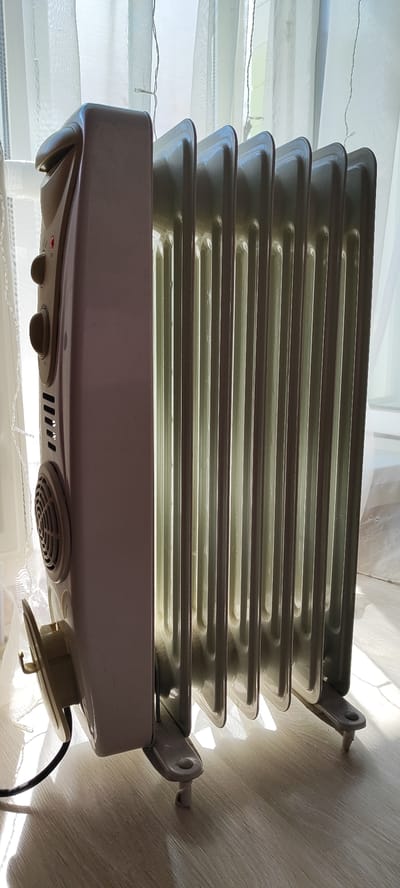An oil-filled radiator is positioned on a light-colored wooden floor in front of a window covered by sheer white curtains. The radiator is beige and has multiple fins. It is plugged into an electrical outlet via a black cord. The radiator has a control panel on its left side with knobs and a red indicator light. The scene is illuminated by sunlight streaming through the window, casting shadows on the floor and the radiator. The location is identified as Snezhinsk, Russia.