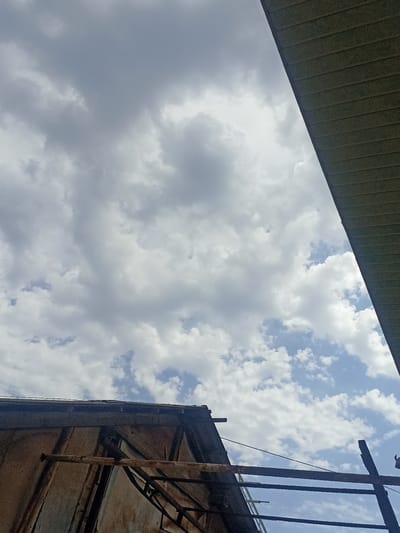 This is a vertical, eye-level medium shot of a partially constructed or dilapidated structure against a cloudy sky. The structure's roof, made of weathered, corrugated metal or wood, occupies the lower left portion of the frame. Exposed metal beams and scaffolding suggest ongoing construction or decay. The sky is filled with a mix of dark and light grey clouds, with patches of blue sky visible. A dark, ridged overhang, possibly part of another building or a large awning, cuts diagonally across the upper right portion of the image. There is no visible text. The lighting suggests it is daytime, possibly late afternoon, with overcast conditions. The scene evokes a sense of a work-in-progress or a forgotten, unfinished space, possibly in an industrial or rural setting. The location context is Chust, Uzbekistan.