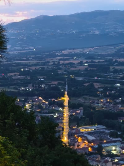 This is a high-angle, vertical shot of a valley at dusk with scattered lights from a town and a highway. The foreground is dominated by dark green trees. In the midground, a road is brightly illuminated by streetlights, leading into a cluster of buildings with lit windows and streetlights. Further back, a highway overpass is visible with faint headlights. The background shows rolling hills dotted with more lights, and wind turbines are silhouetted against a dusky sky with soft pink and blue hues. The scene captures a view of Bojano, Italy, from a higher vantage point.