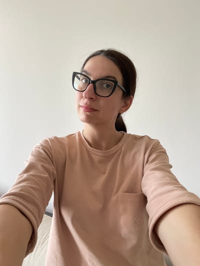 A person is seen from a high angle looking down at the viewer, wearing black-framed glasses and a light pink long-sleeved shirt. The background is a plain white or light-colored surface. No elements indicate a location in Shishmantsi, Bulgaria.