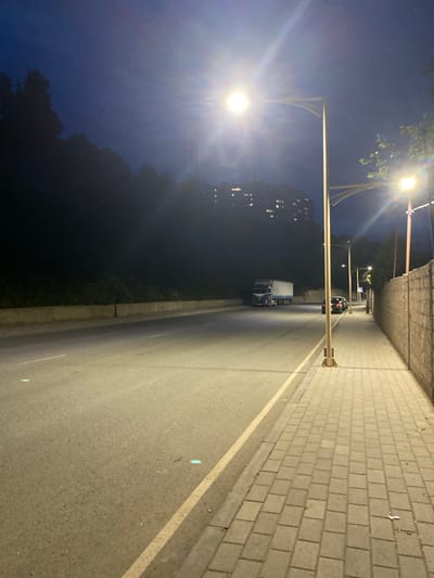The image depicts a street scene at night, likely in Kapan, Armenia. A dimly lit road runs diagonally from the bottom right to the top left of the frame. The road is bordered on the right by a paved sidewalk made of light-colored rectangular bricks. A yellow line marks the edge of the road. On the left side of the road, there is a dark, unlit area, possibly vegetation or a slope, and further back, building lights are visible.

Streetlights with bright, glowing bulbs illuminate the scene. One prominent streetlight on the left casts a strong glare. In the distance on the road, there are two vehicles: a truck with a white trailer and behind it, a car. The sky above is a deep blue, indicating it's nighttime, possibly late evening or early morning. The overall atmosphere is quiet and urban. There is no visible text in the image.
