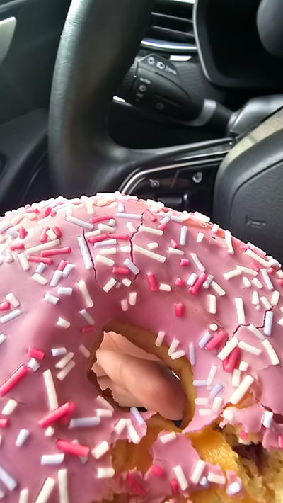 A close-up, vertical shot shows a pink frosted donut with white and pink sprinkles. The donut has cracks in the frosting and appears to be partially eaten. In the background, the steering wheel and dashboard of a car are visible, suggesting the donut is in a vehicle. The steering wheel has controls for lights and wipers. This image was taken in Kamensk-Uralsky, Russia.