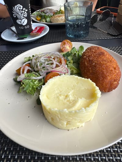 This image shows a close-up view of a meal served on a white plate, set against a dark, woven placemat. The meal consists of a golden-brown, breaded cutlet, a generous portion of mashed potatoes, and a small side salad. The salad includes mixed greens, sliced red onion, and halved cherry tomatoes.
To the left of the plate, a blue glass filled with a clear liquid is visible. In the foreground, a black "TO GO" coffee cup with white decorative elements and coffee bean imagery is placed in a small white dish. This dish also holds a small red packet labeled "Biscott" with a visible biscuit inside. Sunglasses rest on the placemat near the glass.
The lighting suggests it is daytime. The overall setting appears to be an outdoor dining area, possibly a restaurant patio, given the presence of a placemat and tableware. The meal is presented neatly, indicating a dining experience. The image is captured from a top-down perspective, focusing on the food and immediate surroundings. No people are clearly visible, though a hand may be partially present near the coffee cup. The meal appears to be a hearty dish, suitable for lunch or dinner. The environment seems casual and relaxed.