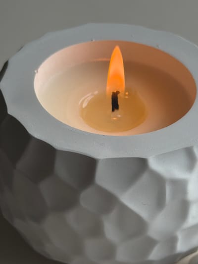 This is a close-up, overhead, and slightly angled shot of a lit candle in a white, geometric-patterned candle holder. The candle's flame is bright orange and yellow, with a dark wick visible at its base. The melted wax forms a pool around the wick. The candle holder's exterior has a textured, faceted surface. The background is a plain, light grey surface. There are no people or other objects visible in the frame. The location in Aespa, Estonia, is not discernible from the image.