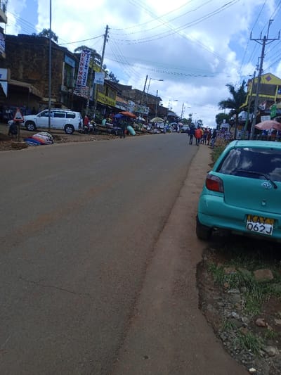 The image displays a street scene in Kangema, Kenya, under a partly cloudy sky. An asphalt road, showing minor surface wear, extends from the foreground into the background, sloping gently uphill.

On the right side of the foreground, a teal Toyota Vitz hatchback is parked on a dirt and gravel shoulder, its rear facing the viewer. The license plate, KAY 062J, is visible. The immediate ground under the vehicle's rear wheel consists of dirt, gravel, and sparse green grass, with a small wet patch present.

The right side of the road features a dirt sidewalk or shoulder populated by multiple pedestrians walking away from the camera. Several individuals are also present on the paved road surface.

On the left side of the road, a continuous line of multi-story commercial buildings borders the street. These structures, appearing to be constructed from stone or painted masonry, house various businesses. At street level, a vibrant market operates, with numerous vendors and customers. Goods, including textiles and what appear to be sacks of produce, are displayed on ground-level stalls, some shaded by colorful umbrellas or awnings. A white SUV is parked on the left near these market stalls, adjacent to a partially visible triangular road sign. Building signage includes a vertical banner for "DENSMAK DRIVING SCHOOL."

Overhead, a dense network of electrical and communication wires spans the width of the street, supported by multiple wooden and metal utility poles. Trees and general vegetation are visible behind the buildings, particularly on the left side, receding into the distance along the street.