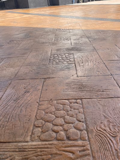The scene depicts an outdoor paved area in Ogbunu, Nigeria, photographed from a low, slightly upward-angled perspective. The foreground and midground consist of a reddish-brown and dark brown stamped concrete pavement, featuring a repeating pattern of rectangular sections with simulated wood grain texture and square sections with simulated cobblestone or river stone texture. The surface appears dry. In the background, a lighter orange-brown paved surface is visible, separated from the foreground pavement by a thin, dark grey or black stripe. Further back and to the left, a dark grey or black wall structure is present. A faint blue object, possibly a bottle or container, is visible near the top right edge of the dark wall. No individuals are present, and no actions or interactions are occurring. The lighting suggests daytime conditions, with shadows cast across parts of the paved surfaces.