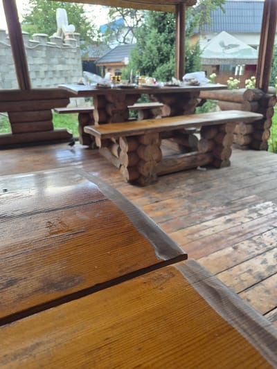 This image depicts an outdoor wooden gazebo, likely a dining or recreational area. The gazebo features rustic wooden tables and benches constructed from logs. In the background, there are trees and buildings, suggesting a semi-rural or park-like setting within the city of Tekeli, Kazakhstan. The focus of the foreground is on the polished wooden surface of a table and the textured wooden floorboards. There are no people clearly visible in the frame, and the scene appears to be empty. The overall impression is one of a natural and weathered outdoor space.