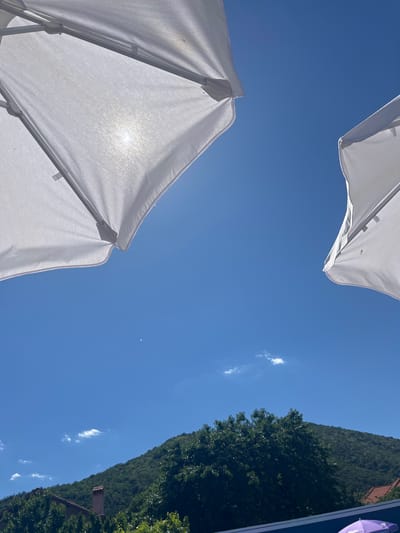 Two white umbrellas are set against a bright blue sky. The sun shines through one of the umbrellas, creating a soft glow. In the distance, a tree-covered hill rises above buildings with red-tiled roofs. A thin crescent moon is visible in the sky. The scene appears to be a sunny day in Varvara, Bulgaria, possibly at a resort or outdoor dining area given the presence of umbrellas.