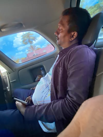 A man with dark hair, a mustache, and closed eyes is asleep in the backseat of a vehicle. He is wearing a dark purple button-up shirt over a white t-shirt with colorful paint-like splatters. His right hand rests on his lap, holding a light-colored smartphone. The interior of the car features a dark gray seat and a dark gray storage compartment in the back, containing a plastic bottle of water and a packet of snacks. The rear window of the car reveals a bright blue sky with white clouds and a reflection of palm trees and a reddish-pink building, suggesting an outdoor, possibly tropical environment. The lighting suggests it is daytime.