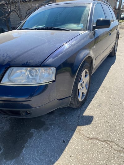 A dark blue Audi A6 C5 Avant (station wagon), heavily coated in a fine layer of dust and pollen, is parked on an asphalt surface under direct sunlight in Dragor, Bulgaria. The vehicle is oriented with its front-left quarter prominently displayed, occupying the majority of the frame. The hood, windshield, and upper body panels show significant dust accumulation, with visible streaks on the dusty paint. The front-left headlight assembly appears clear with minimal hazing, while the lower front bumper consists of a dark grey/black plastic section, revealing an air intake opening. The front-left wheel features a five-spoke silver alloy rim fitted with a tire, visibly marked "POLARISS" on its sidewall. Strong, well-defined shadows from the vehicle are cast onto the grey asphalt, which exhibits minor cracks and dark, localized stains. In the immediate background to the left, a chain-link fence is partially obscured by leafless tree branches, behind which a light-colored building structure is visible. Further down the asphalt path to the right, another light-colored building and a partial view of a distant vehicle are discernible. No individuals or discernible actions are present within the depicted scene.