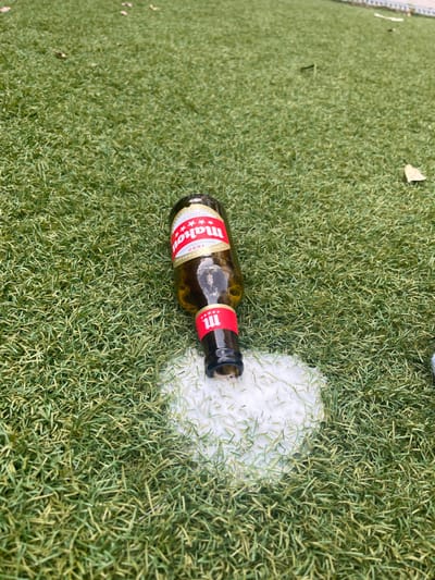 The image shows a partially consumed bottle of "Mahou" beer lying on artificial grass, with a white stain of beer foam around it. The bottle is lying on its side, with the top of the bottle pointing towards the right side of the image. The bottle is partially empty, and the remaining liquid inside is visible. The scene is located in Majadahonda, Spain, at the coordinates 40.469 latitude and -3.857 longitude. There are no people or animals in the image. The background is green artificial grass. The ground appears to be soft. The scene seems to be a public area or park. The image is most likely taken from a high angle perspective, probably from a person's standing height.