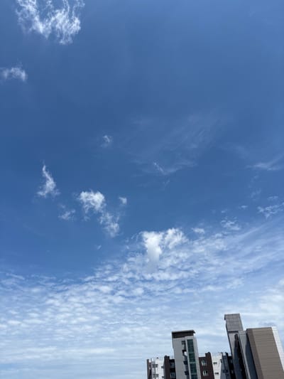 The image shows a clear blue sky with scattered white clouds, and the tops of several modern buildings are visible at the bottom. The buildings appear to be residential or office complexes with contemporary architectural designs, featuring geometric shapes and a mix of gray, brown, and white facades. The sky is vast and bright, with some wispy clouds higher up and more textured, scattered clouds lower down, suggesting a pleasant day. The perspective is upward-looking, emphasizing the sky. The scene is likely from an urban setting in Cheongju-si, South Korea, given the architectural style of the buildings. There are no people visible in the image.