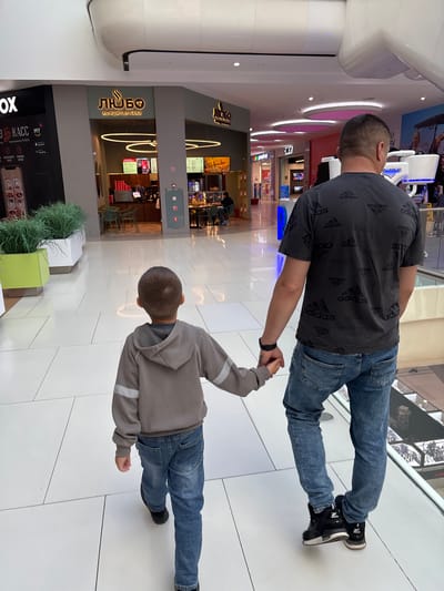In a brightly lit indoor shopping mall, a man and a young boy walk hand-in-hand. The man, seen from behind, wears a dark grey t-shirt with a repeated Adidas logo pattern and light blue distressed jeans. He has a black smartwatch on his left wrist. The boy, also viewed from behind, wears a light grey hooded sweatshirt with reflective silver stripes on the sleeves and blue jeans. They are walking on a polished white tiled floor. To their left, a storefront with a prominent "ЛЮБО" (Lyubo) logo is visible, appearing to be a cafe or confectionery. The environment suggests a modern commercial space with large windows and overhead lighting. The location is identified as Tlustenkhabl, Russia.